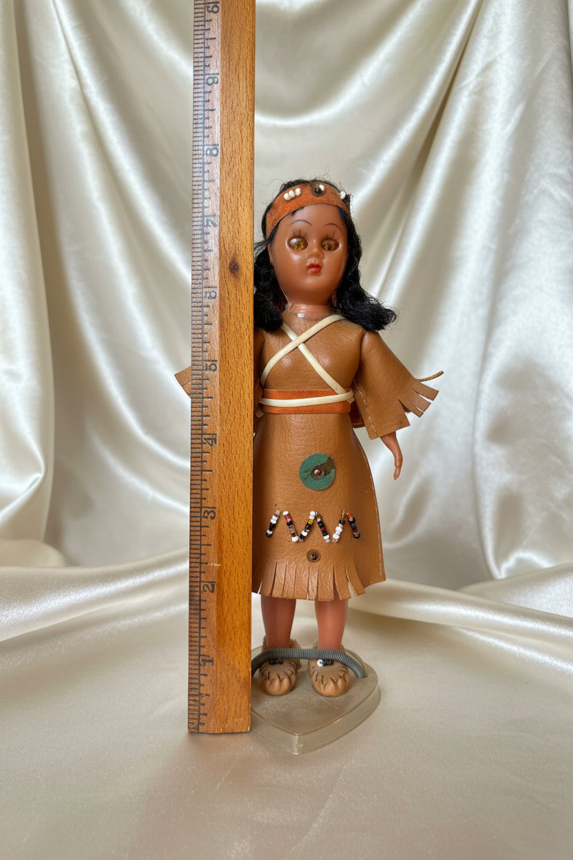 Doll with leather outfit and headdress on a wooden surface with a ruler for scale.