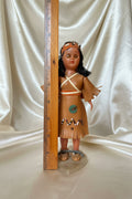 Doll with leather outfit and headdress on a wooden surface with a ruler for scale.
